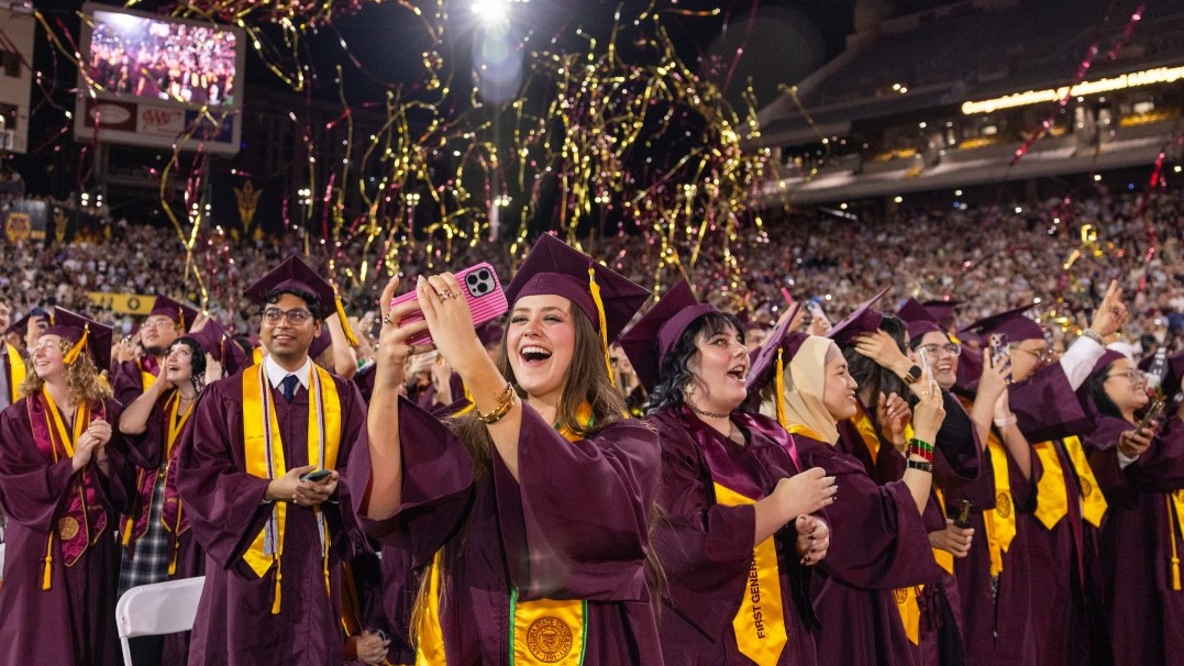ASU students at graduation in caps and gowns with a female student in the foreground taking a photo 