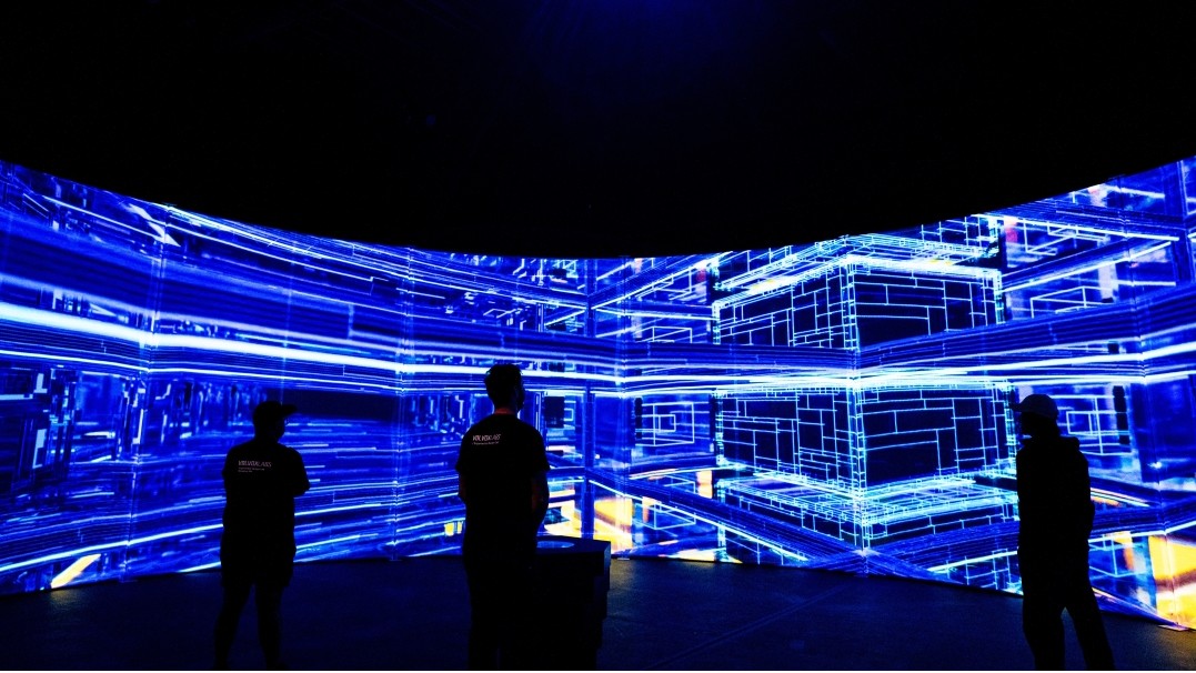 Three people are silhouetted in front of a blue wrap-around screen. 