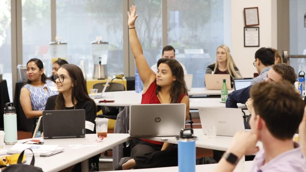 Photo of a classroom with a female student raising her hand and other students looking on 