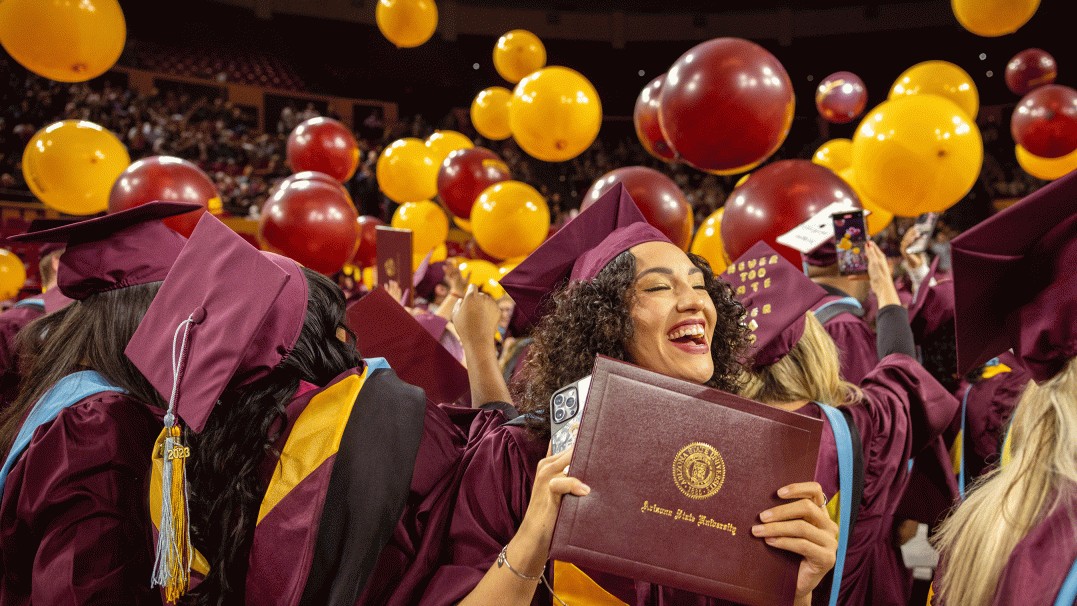 Image of an ASU graduate at commencement smiling and clutching her diploma as balloons rain down on graduates. 