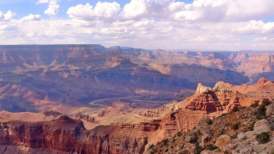Photo of the Grand Canyon with cliffs, red rocks, boulders and puffy clouds in the image. 
