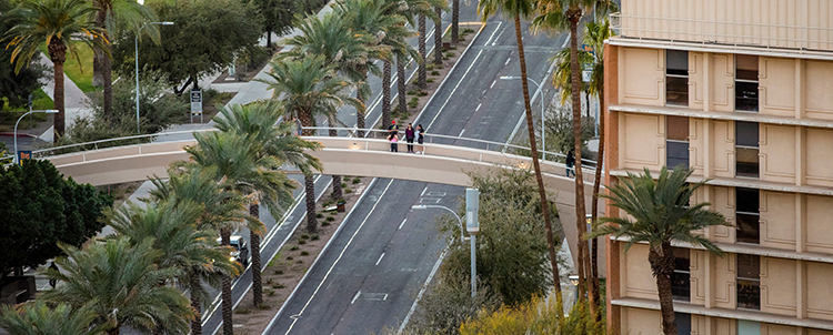 Aerial photo of Tempe campus looking down at a bridge over University Avenue with students on the bridge and a building to the right and palm trees down the center  