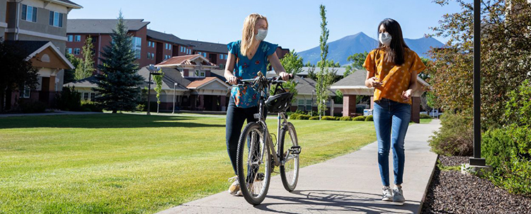 Two female students walking outside on the NAU campus with one walking a bike.