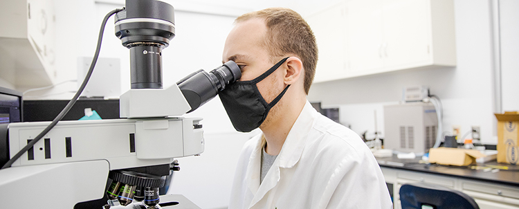 Photo of male researcher in white lab coat and facemask peering through a microscope 