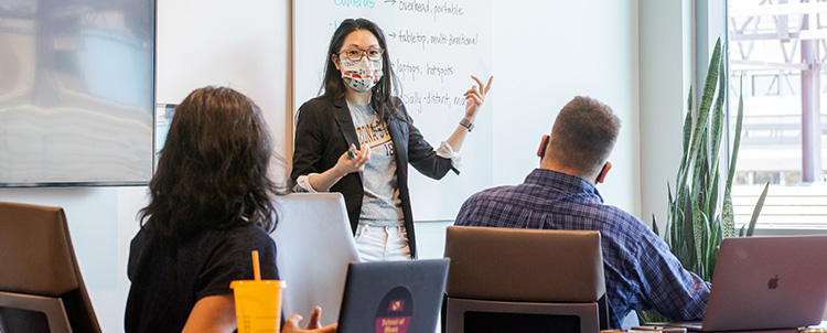 Image of a teacher in front of a classroom with a facemask on with two students with their backs to the camera in the foreground. 
